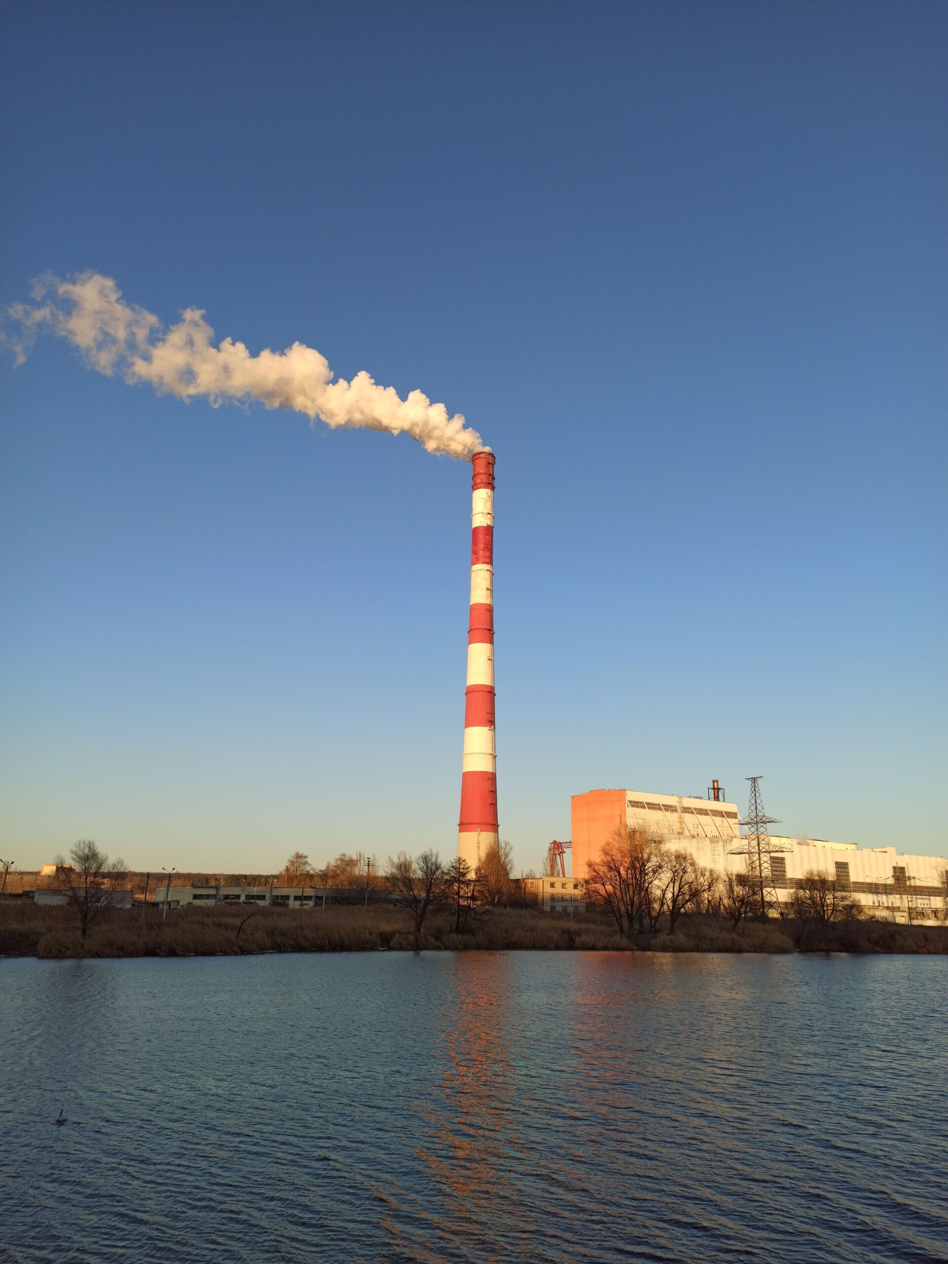 Smoke rises from a power plant chimney near a river in Pisochyn, Ukraine, under a clear sky at dusk.